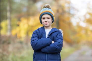 Boy wearing knit hat standing with arms crossed in forest - WPEF04076