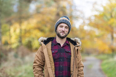 Smiling man wearing knit hat staring while standing at forest - WPEF04077