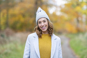 Smiling woman wearing knit hat and jacket staring while standing at forest - WPEF04081