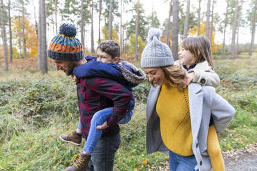 Lächelnde Eltern, die ihre Kinder beim Spaziergang im Wald huckepack nehmen - WPEF04086