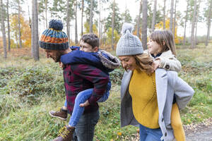 Lächelnde Eltern, die ihre Kinder beim Spaziergang im Wald huckepack nehmen - WPEF04086