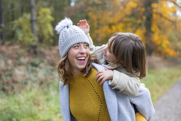 Cheerful mother piggybacking daughter while standing at forest - WPEF04087