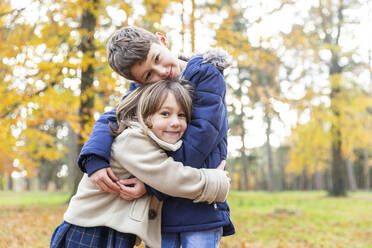 Smiling brother and sister embracing while standing in forest - WPEF04135