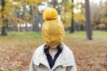 Girl wearing jacket covering face with knit hat while standing at forest - WPEF04161