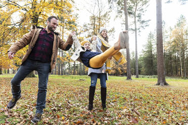 Smiling girl swinging while holding hands of parents standing in forest - WPEF04165