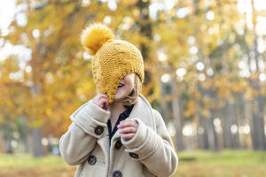 Playful girl covering face with knit hat while standing at forest - WPEF04174