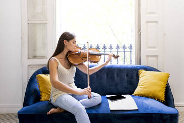 Musikerin spielt zu Hause auf einem Sofa mit einem Tablet Bratsche. - CAVF93379