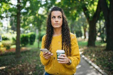 Frau mit Smartphone und Plastikbecher mit Kaffee im Park. - CAVF93396