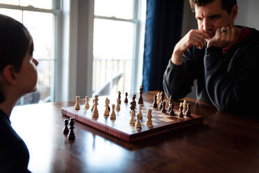 Vater und Sohn sitzen an einem Tisch in einem Haus und spielen eine Partie Schach. - CAVF93408