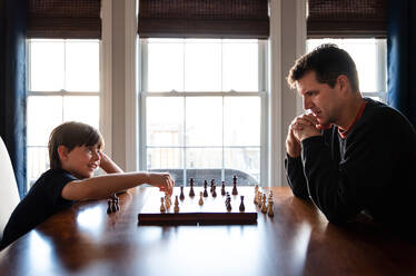 Vater und Sohn sitzen an einem Tisch in einem Haus und spielen eine Partie Schach. - CAVF93410