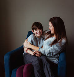 Happy woman hugging her smiling son as he sits in her lap on a chair. - CAVF93411