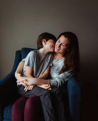 Happy woman hugging her son as he sits in her lap on a chair. - CAVF93412