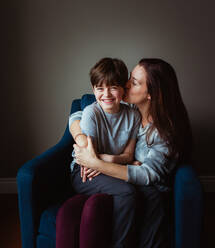 Woman hugging and kissing her son as he sits in her lap on a chair. - CAVF93413