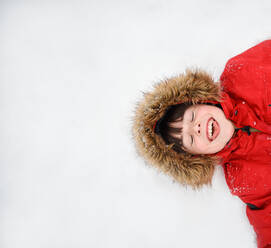 Overhead des glücklichen Jungen im roten Mantel mit pelziger Kapuze, der im Schnee liegt. - CAVF93414