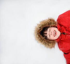 Overhead of happy boy in red coat with furry hood laying in the snow. - CAVF93414