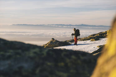 Young man wearing cap and looking at sun rising above horizon, against clouds, Gredos, Spain - CAVF93432