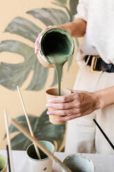 Abgeschnittene unerkennbare Künstlerin mit Maske, die Farbe in einen Becher gießt und Farben mischt, um Monstera-Blätter an der Wand zu Hause zu gestalten - ADSF20748