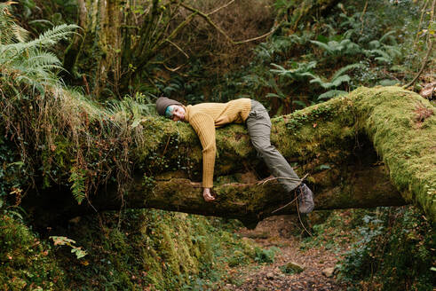 Side view of female explorer lying on tree trunk and having break during trekking in mossy forest - ADSF20752