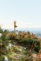 Back view of hiker standing in Vrksasana on rock and practicing yoga in mountains during trekking at sunset - ADSF20757