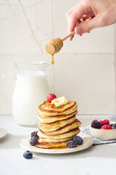 Pouring honey on a stack of fluffy breakfast pancakes with berries and butter - ADSF20766