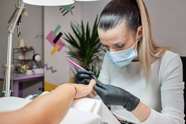 Professional master in protective mask and gloves using machine while polishing nails of female customer in manicure salon - ADSF20772