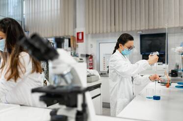 Professionelle Wissenschaftlerin im weißen Kittel und mit Schutzmaske führt ein chemisches Experiment mit einer blauen Flüssigkeit in einem modernen Labor durch - ADSF20786