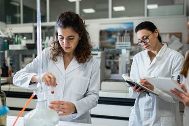 Konzentrierte Wissenschaftlerin, die ein chemisches Experiment mit einer Flüssigkeit in einem Glaskolben durchführt, während ihre Kollegen während der Forschung im Labor die Ergebnisse aufschreiben - ADSF20790
