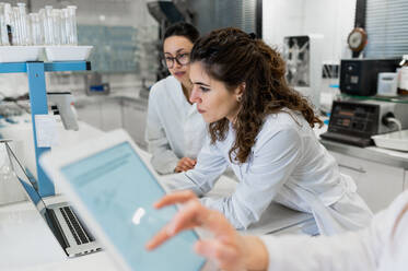 Junge Pharmazieforscherinnen versammeln sich an einem Tisch mit einem Laptop mit Diagrammen und diskutieren Forschungsergebnisse, während sie gemeinsam in einem wissenschaftlichen Labor arbeiten - ADSF20794
