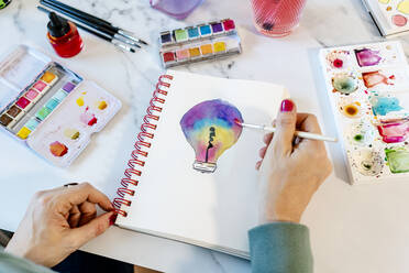 Woman painting light bulb with watercolors on spiral notebook at table - GEMF04671