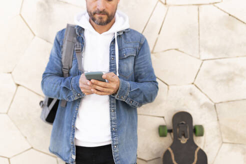 Mann mit Skateboard, der sein Smartphone benutzt und sich an die Wand lehnt - JPTF00657