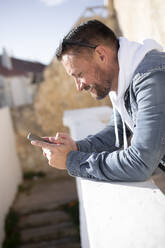 Smiling mature man using smart phone while leaning on retaining wall - JPTF00660