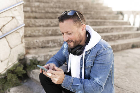 Mann mit Kopfhörern, der sein Smartphone benutzt, während er auf einer Treppe sitzt - JPTF00661