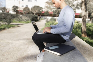 Mature businessman using laptop at while sitting on concrete bench public park - JPTF00664