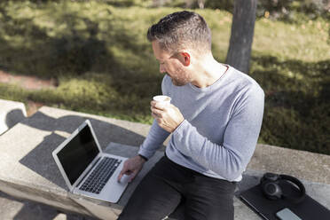 Älterer Geschäftsmann mit Einweg-Kaffeebecher, der einen Laptop benutzt, während er auf einer Betonbank in einem öffentlichen Park sitzt - JPTF00666