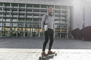 Männlicher Pendler mit Akten, der auf einem Skateboard vor einem Gebäude steht und wegschaut - JPTF00674