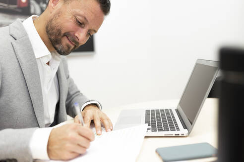 Male entrepreneur with laptop writing on paper at desk in office - JPTF00678