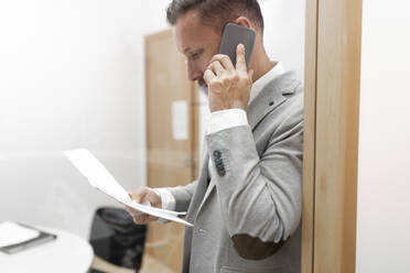 Geschäftsmann mit Dokumenten im Gespräch über Smartphone im Büro - JPTF00680