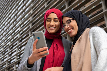 Niedriger Winkel der arabischen besten Freundinnen in Hidschab, die auf der Straße stehen und im Internet auf dem Smartphone surfen, während sie lachen und Spaß haben - ADSF20821