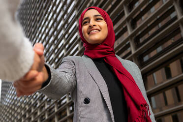 From below of Cheerful Arab female entrepreneur shaking hand of crop business partner while standing in city - ADSF20823