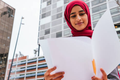 Niedriger Winkel einer arabischen Studentin mit traditionellem Kopftuch, die auf der Straße sitzt und Unterlagen für ihre Hausaufgaben liest - ADSF20825