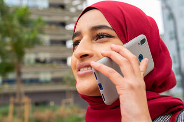 Niedriger Winkel der positiven ethnischen Frau im Hidschab, die auf der Straße steht und mit ihrem Smartphone spricht und das Wochenende genießt - ADSF20827