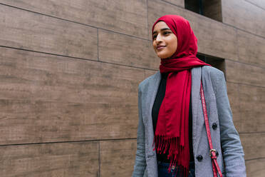 Zufriedene junge arabische Frau mit traditionellem Hidschab und Freizeitkleidung, die eine Straße entlanggeht und nach vorne schaut - ADSF20831