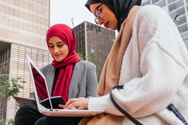 Inhalt Arabische Studentinnen mit Kopftuch sitzen auf einer Bank im grünen Garten des Campus und bereiten sich auf Prüfungen vor, während sie Notizen lesen und einen Laptop benutzen - ADSF20840