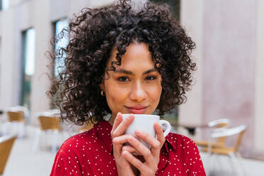Glückliche junge ethnische Frau mit lockigem Haar, die eine Tasse aromatischen Kaffee genießt und in die Kamera schaut, während sie sich in einem Café im Freien ausruht - ADSF20849