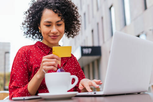 Happy young ethnic female with credit card in hand using laptop and doing online purchase on internet shop while sitting at table in outdoor cafe - ADSF20850