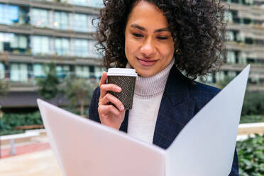Fröhliche afroamerikanische Unternehmerin mit einer Tasse Kaffee zum Mitnehmen, die Dokumente liest, während sie in der Stadt steht - ADSF20852