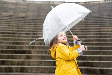 Playful kid in yellow raincoat and with wet umbrella standing on street enjoying rainy weather in city - ADSF20859