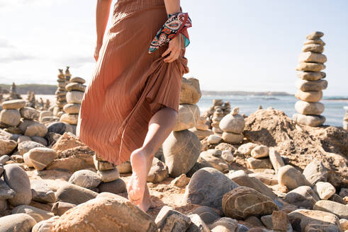 Cropped unerkennbar junge weibliche Reisende in stilvollem Outfit zu Fuß barfuß auf Sandstrand inmitten von Steinen während des Urlaubs in Algarve - ADSF20862