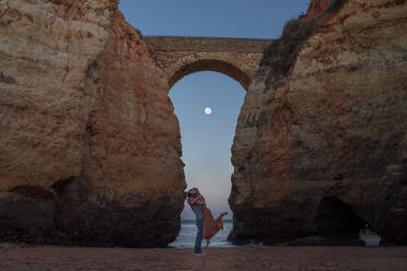 Rückenansicht eines nicht erkennbaren männlichen Reisenden, der eine glückliche Freundin auf der Schulter trägt, während er sich gemeinsam am berühmten Sandstrand Praia de Estudantes entspannt und das Meer gegen den wolkenlosen Abendhimmel bewundert - ADSF20867