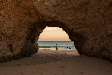 Seitenansicht einer nicht erkennbaren Frau in Freizeitkleidung und Hut, die auf Sand am Eingang einer Höhle in der Nähe des Meeres bei Sonnenuntergang in den Benagil-Höhlen an der Algarve, Portugal, steht - ADSF20869